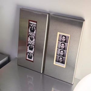 May include: Two rectangular metal photo frames. One frame has a red border and contains a strip of black and white photos of a group of people. The other frame has a beige border and contains a strip of black and white photos of a person.