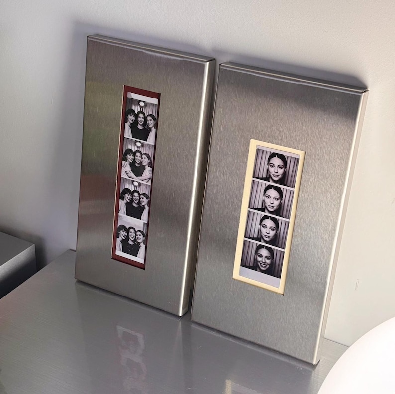 May include: Two rectangular metal photo frames. One frame has a red border and contains a strip of black and white photos of a group of people. The other frame has a beige border and contains a strip of black and white photos of a person.