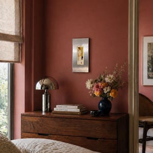 May include: A bedroom scene featuring a dark wooden dresser with a silver lamp and books. A blue vase holds a bouquet of flowers. A framed artwork hangs on the reddish-brown wall. A bed with a beige comforter is in the foreground.