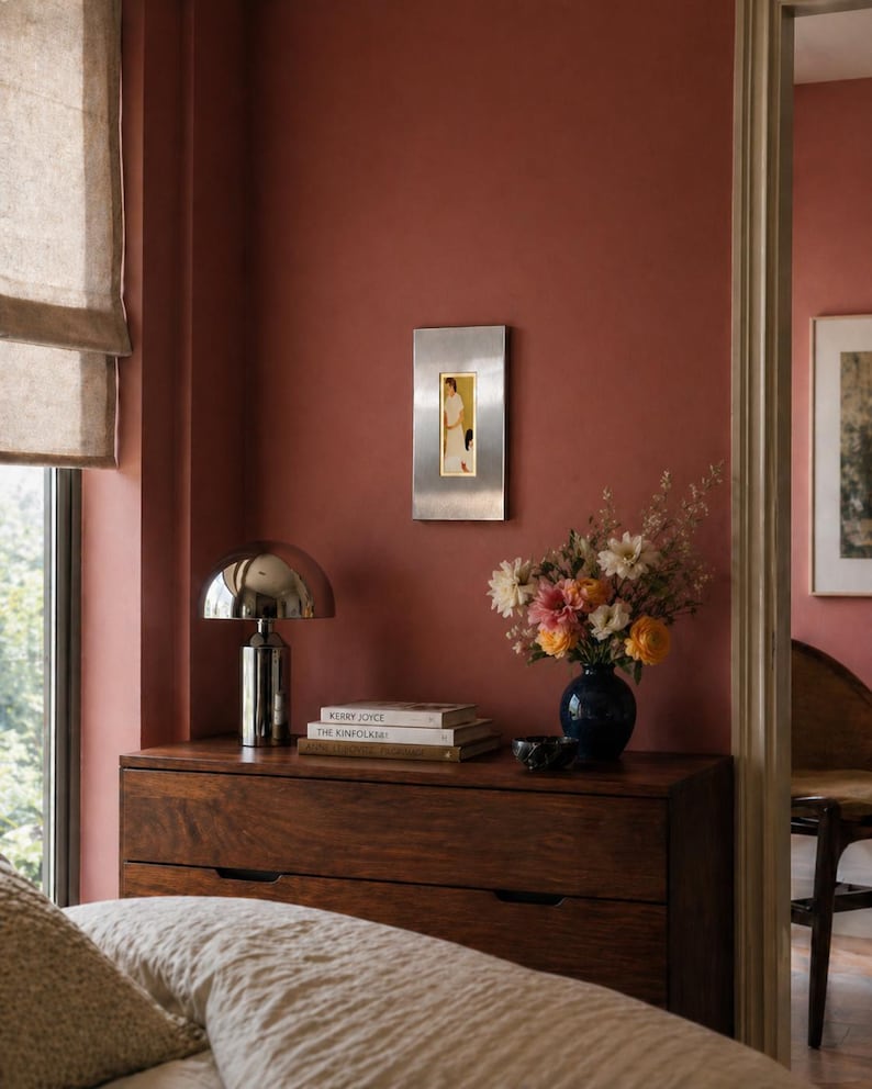 May include: A bedroom scene featuring a dark wooden dresser with a silver lamp and books. A blue vase holds a bouquet of flowers. A framed artwork hangs on the reddish-brown wall. A bed with a beige comforter is in the foreground.