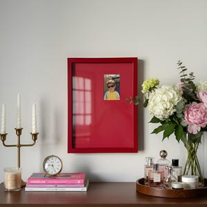 May include: A vibrant red picture frame, displaying a child's photo, is mounted on a white wall. A gold candelabra with white candles, books, and a small clock are on a wooden surface. A bouquet of flowers and perfume bottles are also on the surface.