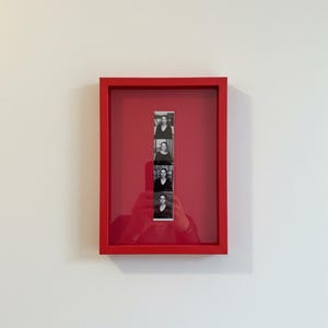 May include: A red framed art piece with a red background. The artwork features a strip of four black and white portrait photographs. The frame is rectangular and mounted on a white wall.