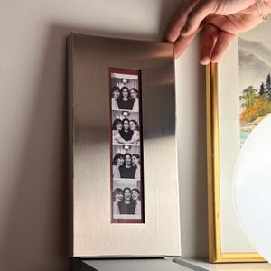 Puede incluir: Un marco de fotos rectangular de metal cepillado con una abertura vertical estrecha. En el interior, se ven una serie de imágenes en blanco y negro al estilo de fotomatón de personas sonrientes. El marco está siendo sostenido por una mano.