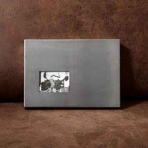 May include: A rectangular, brushed silver-toned metal photo frame with a smooth, solid surface. A small, centered rectangular cutout displays a black and white photograph of two people standing in an interior space, captured in a candid pose.