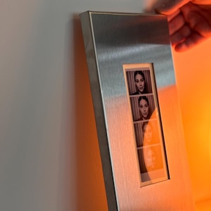 May include: A rectangular, brushed metal photo frame with a vertical opening displaying four black and white photo booth style images of a person. The frame is held by a hand against a white wall.
