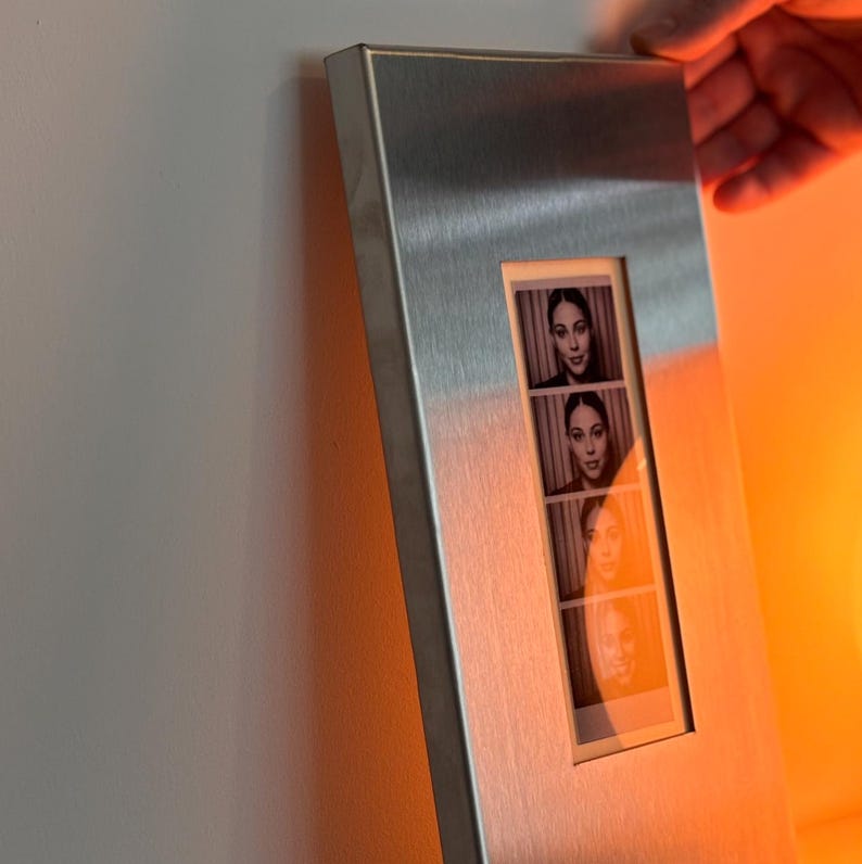 May include: A rectangular, brushed metal photo frame with a vertical opening displaying four black and white photo booth style images of a person. The frame is held by a hand against a white wall.