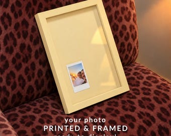 May include: A light yellow picture frame containing a small instant photograph of two people at sunset. The text below the frame reads: your photo PRINTED & FRAMED ready to display! The frame has a smooth, matte finish and a rectangular shape.