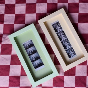Pode incluir: Dois porta-retratos retangulares, um em verde claro e outro em creme, cada um contendo uma tira de fotos vertical em preto e branco. As molduras têm um acabamento liso e um design profundo, exibindo retratos em quatro quadros distintos.