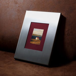 May include: A rectangular picture frame with a brushed silver metal finish and a deep burgundy mount. The frame contains a small landscape print depicting a white house with a red roof situated in a grassy field near a body of water.