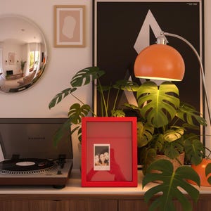 Könnte beinhalten: Ein roter Bilderrahmen mit einem Foto von drei Personen steht auf einer Holzoberfläche. Ein Plattenspieler und eine große orangefarbene Lampe sind ebenfalls zu sehen. Der Hintergrund umfasst ein schwarz-weißes Poster, einen runden Spiegel und Topfpflanzen.