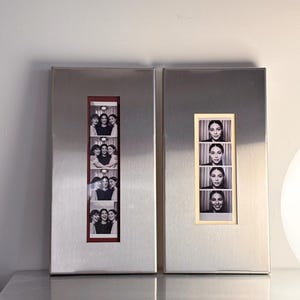 May include: Two rectangular, brushed metal photo frames. One frame displays a series of black and white photo booth images of three women. The other frame shows a series of black and white photo booth images of a woman.
