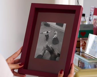 Könnte beinhalten: Ein tiefburgunderroter, rechteckiger Bilderrahmen mit matter Oberfläche und passendem Passepartout. Der Rahmen enthält ein Schwarz-Weiß-Foto, das zwei Personen beim Tanzen im Freien aus einer erhöhten Perspektive zeigt.