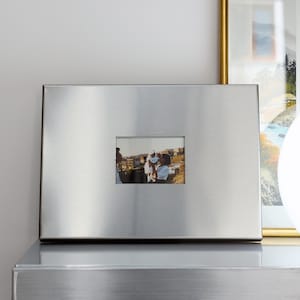 May include: A rectangular, brushed silver metal picture frame with a wide, flat border. The frame features a centered, smaller rectangular cutout that displays a photograph of two people holding a baby outdoors in a sunny, hilly landscape.