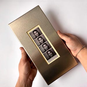 May include: A rectangular, brushed metal photo frame holds a strip of four black and white photo booth images. The frame has a gold-toned finish and a rectangular cutout to display the photos. The photos show a woman with different expressions.