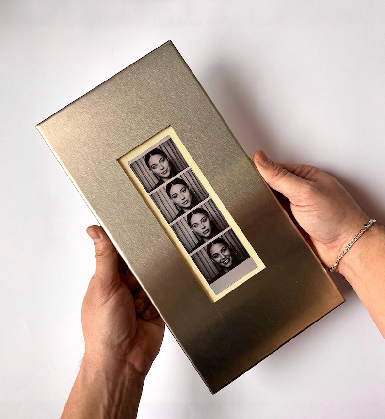 May include: A rectangular, brushed metal photo frame holds a strip of four black and white photo booth images. The frame has a gold-toned finish and a rectangular cutout to display the photos. The photos show a woman with different expressions.