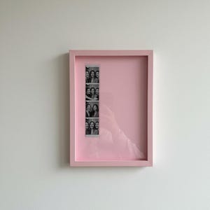 May include: A pink rectangular picture frame with a strip of black and white photo booth pictures. The frame is mounted on a white wall. The frame has a pink border and a pink interior.