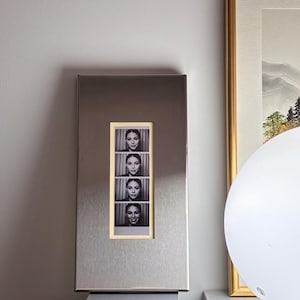 May include: A silver metal photo frame with a rectangular window displaying four black and white photo booth style images of a person. The frame is mounted on a gray wall.