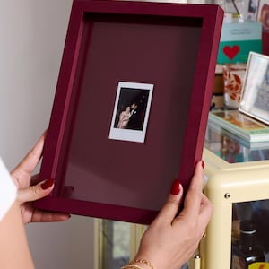 May include: A deep burgundy rectangular picture frame featuring a matching mat board with a centred cutout. A small, vertical instant film photograph showing two people is displayed within the cutout, protected by a clear glass front panel.