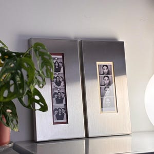 May include: Two rectangular silver photo frames, each holding a strip of black and white photos. One frame displays images of three women, while the other shows a woman's portrait. The frames are displayed on a gray surface.