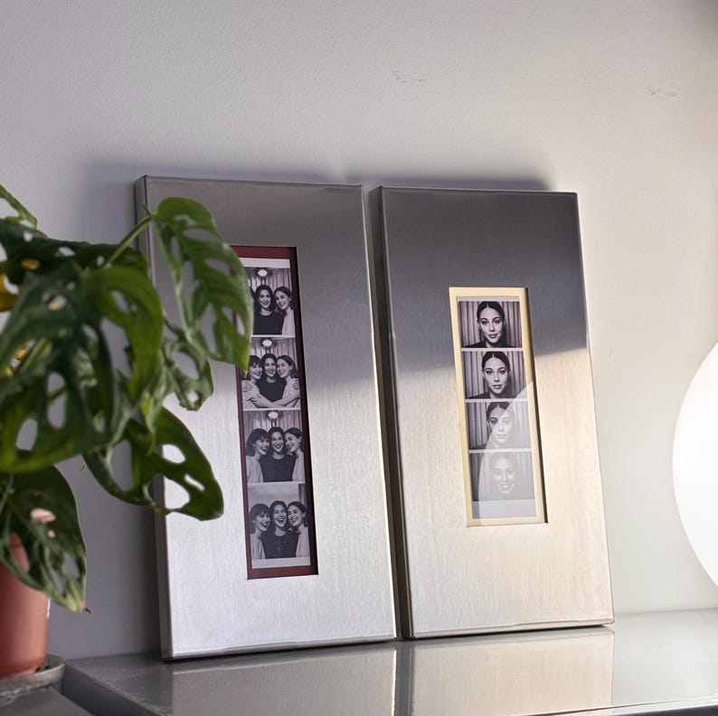 May include: Two rectangular silver photo frames, each holding a strip of black and white photos. One frame displays images of three women, while the other shows a woman's portrait. The frames are displayed on a gray surface.