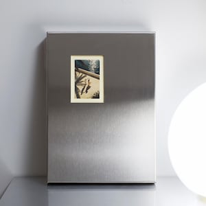 Könnte beinhalten: Ein rechteckiges Fotoalbum mit einem Einband aus gebürstetem silberfarbenem Metall. Auf der Vorderseite befindet sich ein mittiger, vertikaler rechteckiger Ausschnitt, der ein kleines Foto von zwei Personen an einem sonnigen Strand zeigt.