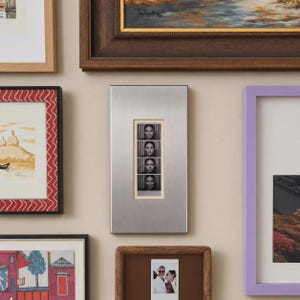 May include: A rectangular silver-toned metal picture frame featuring a vertical cutout that displays a black and white photo strip containing four sequential portraits of the same person with varying facial expressions, centered within the frame.
