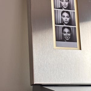 May include: A black and white photo strip featuring three portraits of a person with dark hair and a neutral expression. The strip is set against a metallic, possibly stainless steel, background. The portraits show the person in different poses.