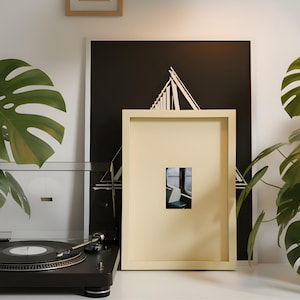 May include: A black turntable with a record is positioned in front of a framed print. The print features a small image of a train interior within a larger cream-colored frame. A black and white graphic print is behind the cream frame.