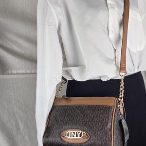 Puede incluir: Bolso bandolera marrón y beige con correa de cadena dorada. El bolso presenta un patrón repetitivo y un emblema ovalado dorado que dice "DNY". El bolso se muestra en un maniquí con una camisa blanca abotonada y una falda negra.