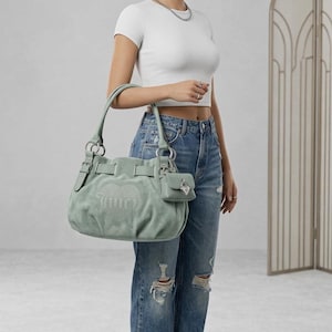 May include: A sage green suede shoulder bag featuring a large embossed heart logo with the word Juicy in the center. The bag includes silver-tone hardware, buckled side straps, and a small matching detachable pouch with a heart-shaped padlock charm.