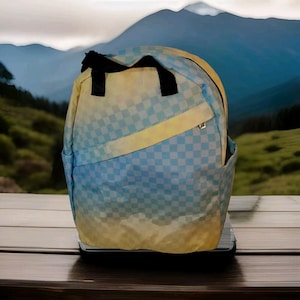 May include: A backpack with a blue and yellow checkered pattern. The backpack has a black handle and a front pocket. The background shows a mountain landscape.