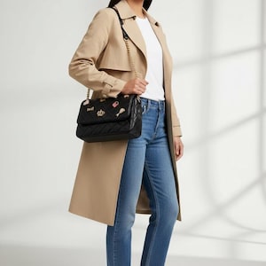 Op de afbeelding: Een zwarte gewatteerde handtas met een gouden kettingriem en decoratieve patches. De persoon draagt een beige trenchcoat, een wit shirt, een blauwe jeans en bruine loafers.
