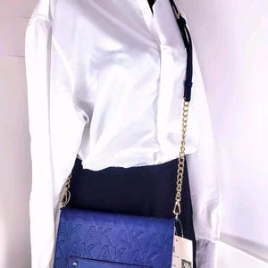 Puede incluir: Un bolso bandolera azul marino con una correa de cadena dorada. El bolso presenta un patrón en relieve de las letras "AKAK" y una pequeña etiqueta. El bolso cuelga de una camisa blanca abotonada y una falda negra.