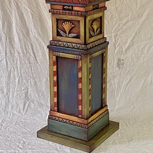 May include: A hand-painted wooden pedestal with a square top and a multi-layered design. The pedestal is painted in shades of green, yellow, red, and blue, with intricate details and geometric patterns.