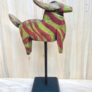 May include: A hand-carved wooden sculpture of a bull, painted with red and green stripes. The bull has large horns and a tail. It is mounted on a black metal stand with a square base. The sculpture is approximately 8 inches tall.
