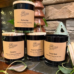 May include: Four black glass candles with wooden lids and labels. The labels read "Barbershop", "Campfire", "Pocket of Sunshine", and "Forest Bath". Each candle is labeled "RB Apothecary" and lists the scent and that it is 100% soy.