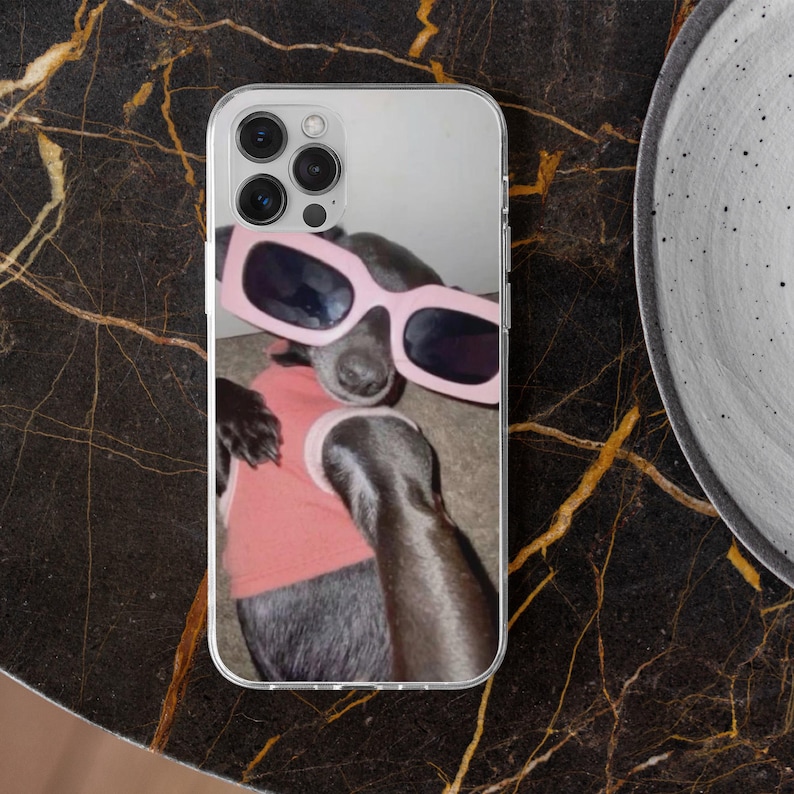 May include: Clear phone case featuring a dog wearing pink sunglasses and a pink shirt. The dog is gray with a black nose and is lying down. The phone case has a clear border and a camera bump.
