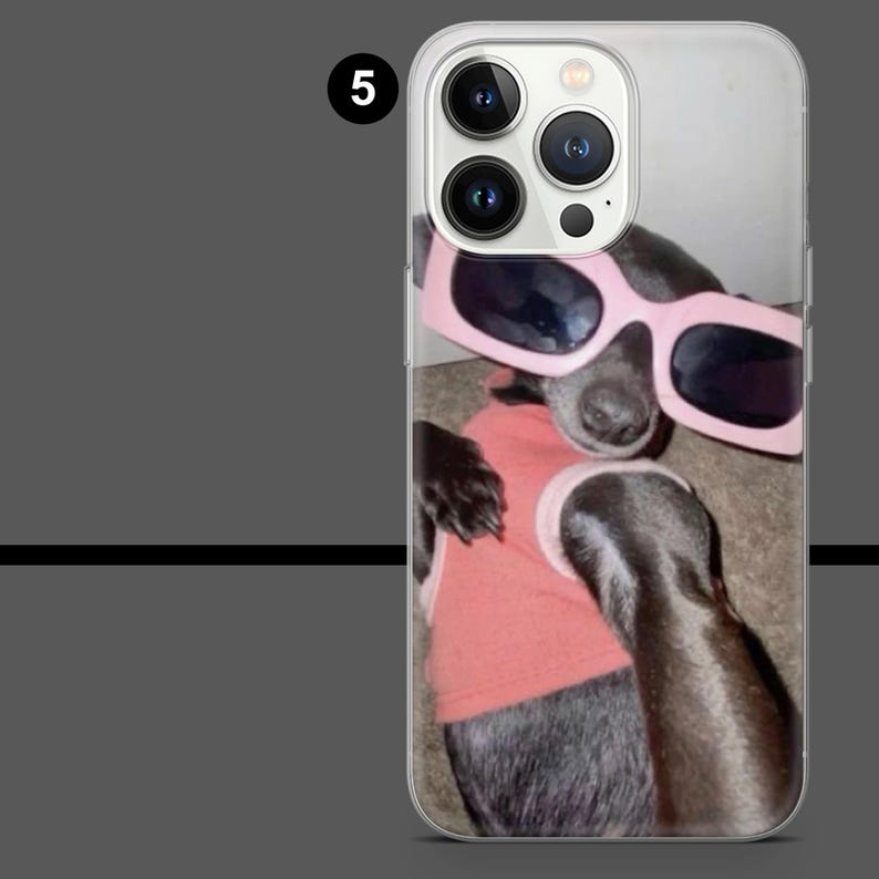 May include: Clear phone case featuring a black dog wearing pink sunglasses and a pink shirt. The dog is lying down, with its paws visible. The case has a clear border and a camera cutout.