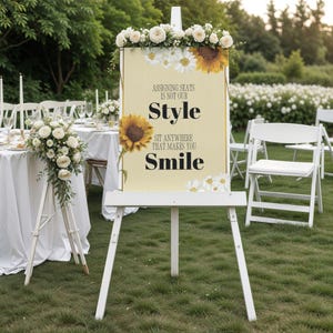 May include: A white easel displays a sign with the text "Assigning seats is not our style. Sit anywhere that makes you smile." The sign is decorated with sunflowers and daisies. The scene is set outdoors with white chairs and tables.