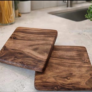 May include: Two rectangular wooden cutting boards with rounded corners. The boards are made of dark brown wood with a prominent grain pattern. They are displayed on a light-colored countertop, suggesting a kitchen setting. The boards are likely used for food preparation.