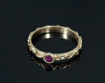 May include: A delicate gold band ring with a single, small, round, purple gemstone set in the center. The band is decorated with a repeating pattern of small, curved lines.