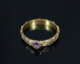 May include: A gold band ring with a purple gemstone set in the center. The band is decorated with a swirling pattern.