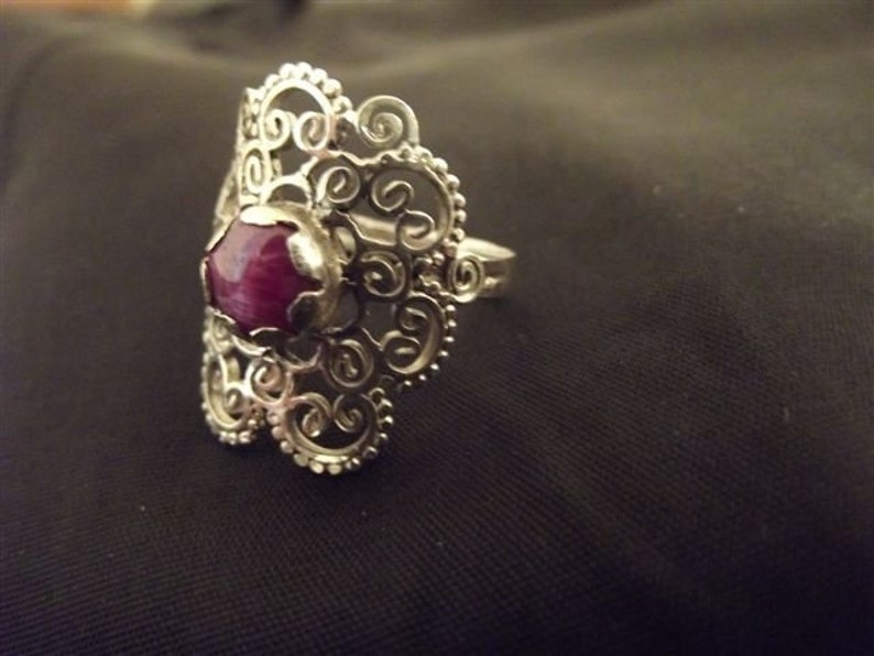 May include: A silver filigree ring with a large, round, dark pink gemstone.