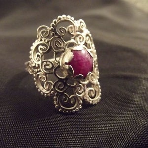 May include: A silver filigree ring with a large, round, dark pink gemstone.