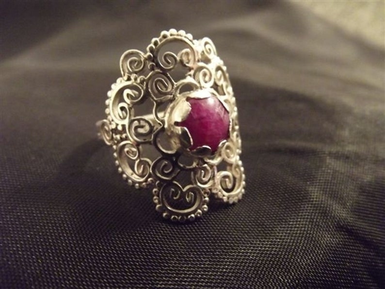 May include: A silver filigree ring with a large, round, dark pink gemstone.