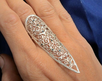 May include: A silver filigree ring with a pointed design and small blue gemstones. The ring is worn on a finger.