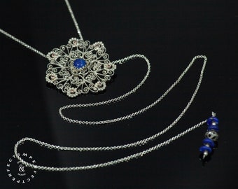 May include: A silver necklace with a large, intricate pendant featuring a blue gemstone. The pendant is made of delicate, swirling wire and has a floral design. The necklace also has a long chain with small blue beads at the end.