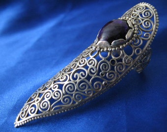 May include: A silver filigree ring with a large, dark purple gemstone. The ring is designed to be worn on the middle finger and has a pointed, elongated shape.