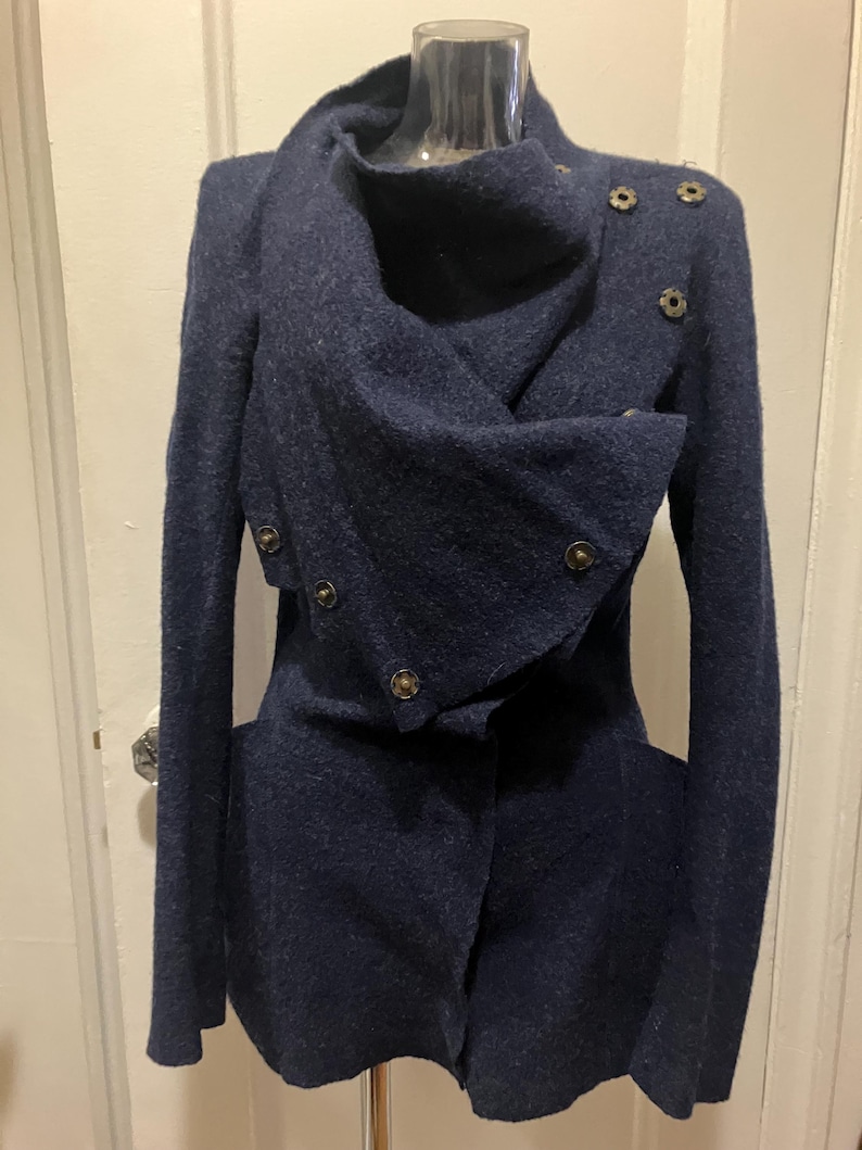 Puede incluir: Un abrigo de lana azul marino con un dise&ntilde;o asim&eacute;trico. El abrigo presenta un cuello drapeado, mangas largas y varios botones decorativos. El abrigo se exhibe en un maniqu&iacute;.