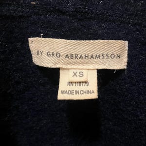 Puede incluir: Primer plano de una etiqueta de prenda azul marino. La etiqueta dice "BY GRO ABRAHAMSSON" y "XS". Debajo de la talla, la etiqueta indica "RN 118779" y "MADE IN CHINA". La prenda parece ser un su&eacute;ter o c&aacute;rdigan.